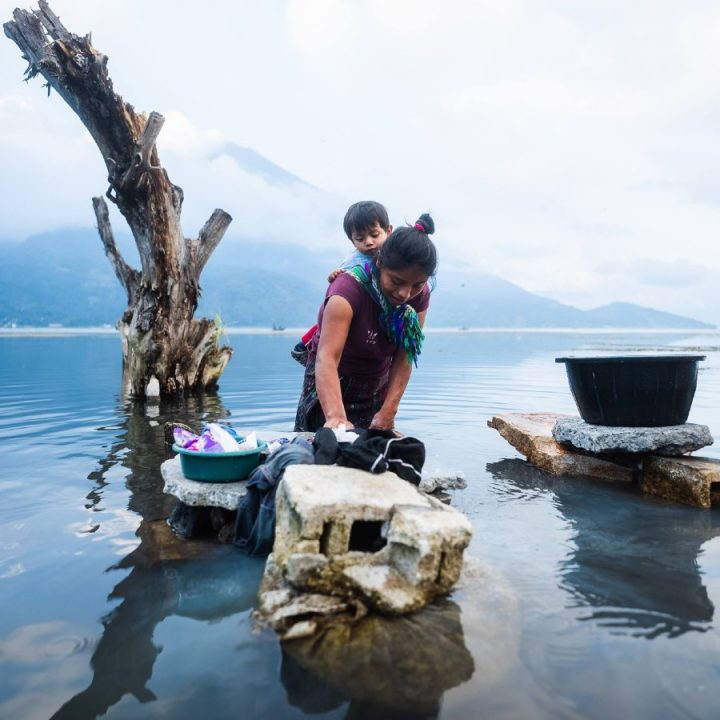 Young woman holding young child on her back as she washes clothes in a lake